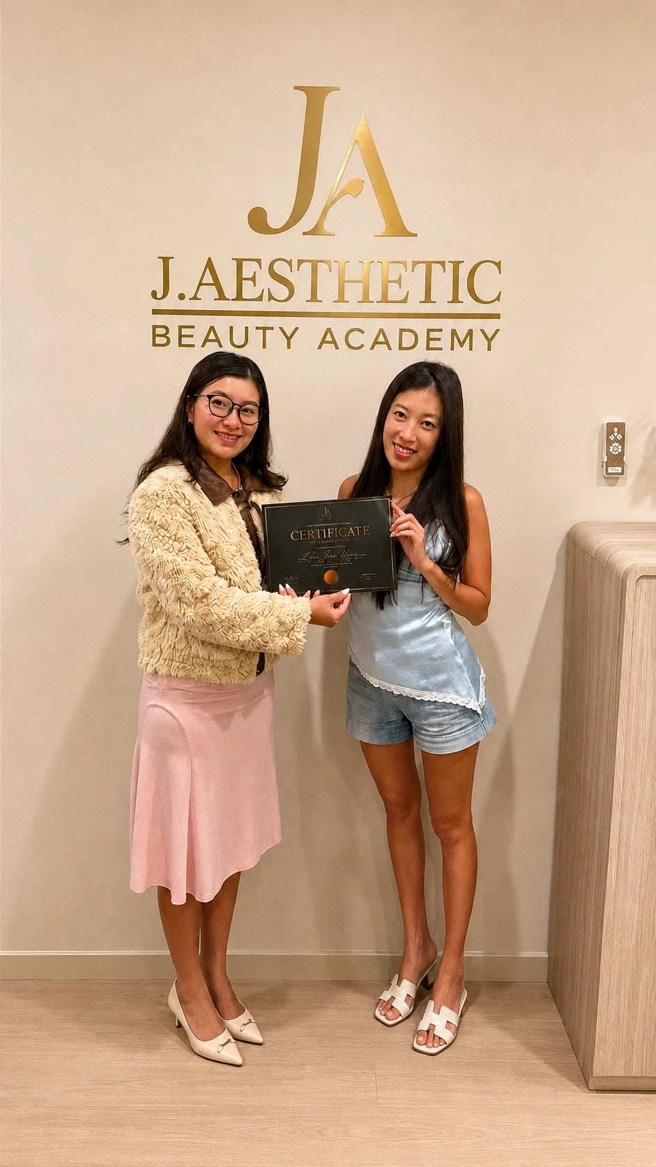 Two women standing indoors in front of a beige wall with a large gold 'JA AESTHETIC BEAUTY ACADEMY' logo. The woman on the left wears a cream textured cardigan and pink skirt, and the woman on the right wears a light blue satin top and denim shorts.