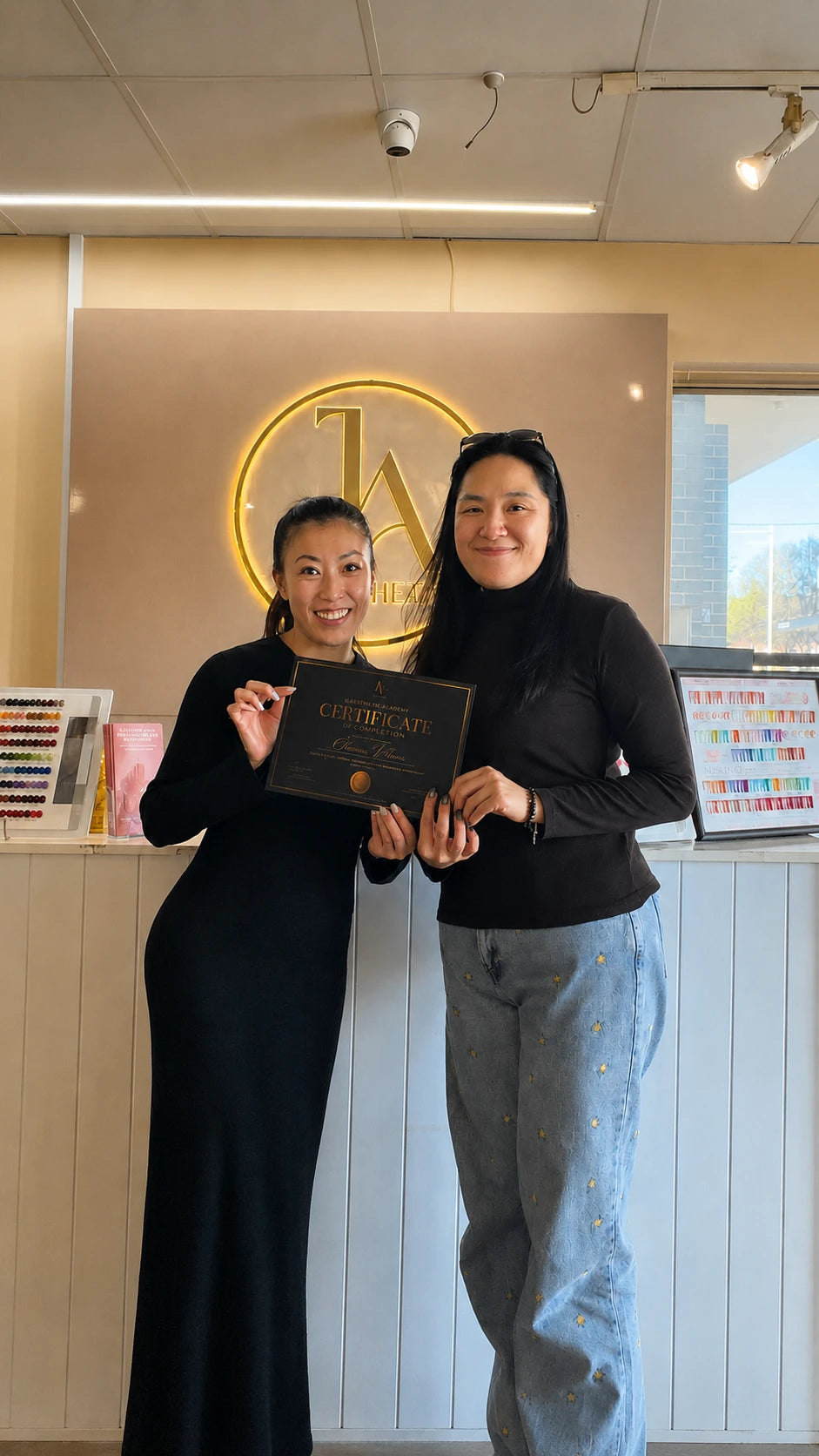 Two women standing indoors in front of a beige wall with a large gold circular logo. One woman wears a long black dress and holds a certificate plaque, while the other wears a black long-sleeve top and light blue jeans.