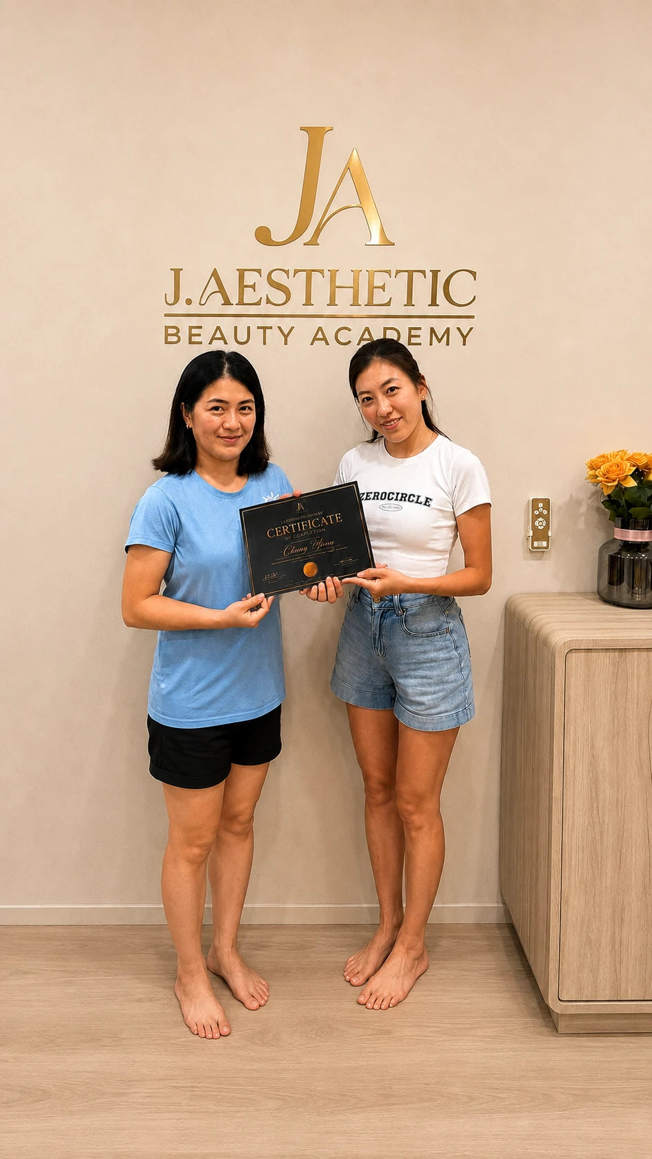 Two women standing barefoot in a beauty academy interior, holding a black certificate. The wall behind them displays the gold 'J.
