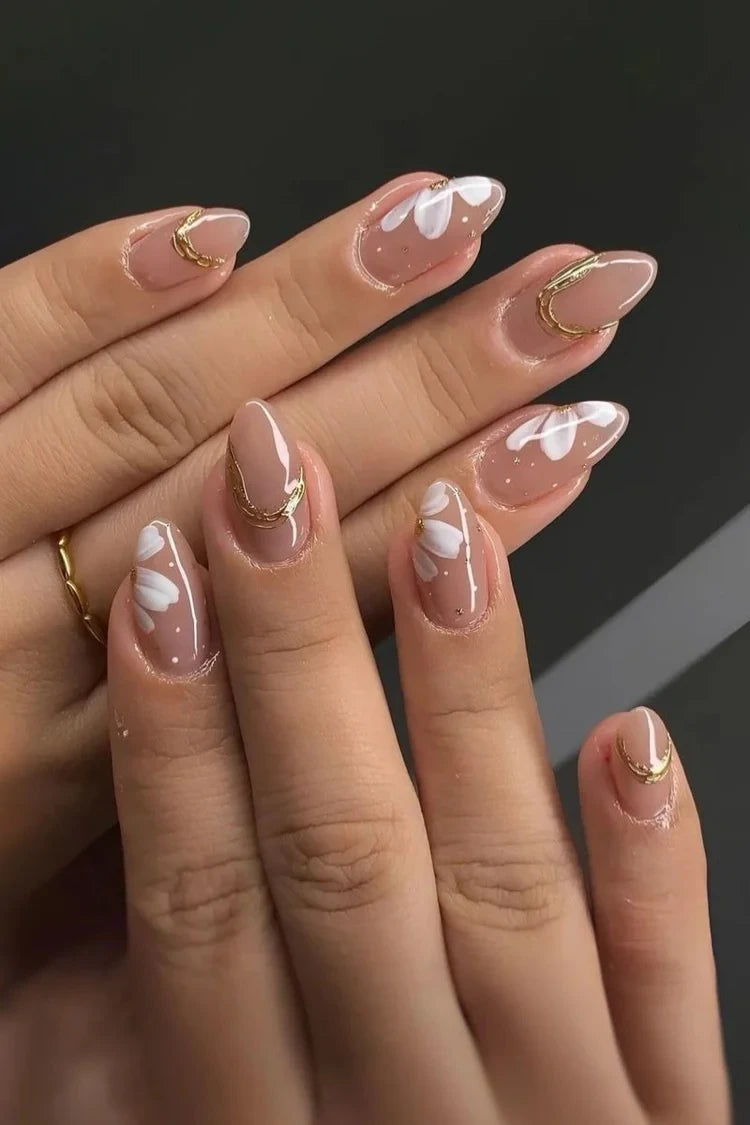Close-up of almond-shaped nude nails with white floral nail art and gold accent details